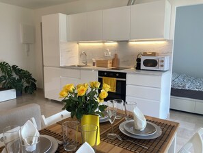 Una cocina moderna con armarios blancos, un microondas y un lavamanos. Hay una mesa de comedor con un jarrón de flores amarillas y un mantel individual.