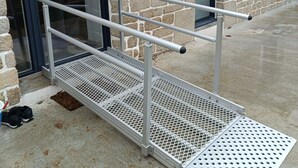 Een metalen hellingbaan die leidt naar de ingang van een gebouw met stenen muren en een glazen deur.