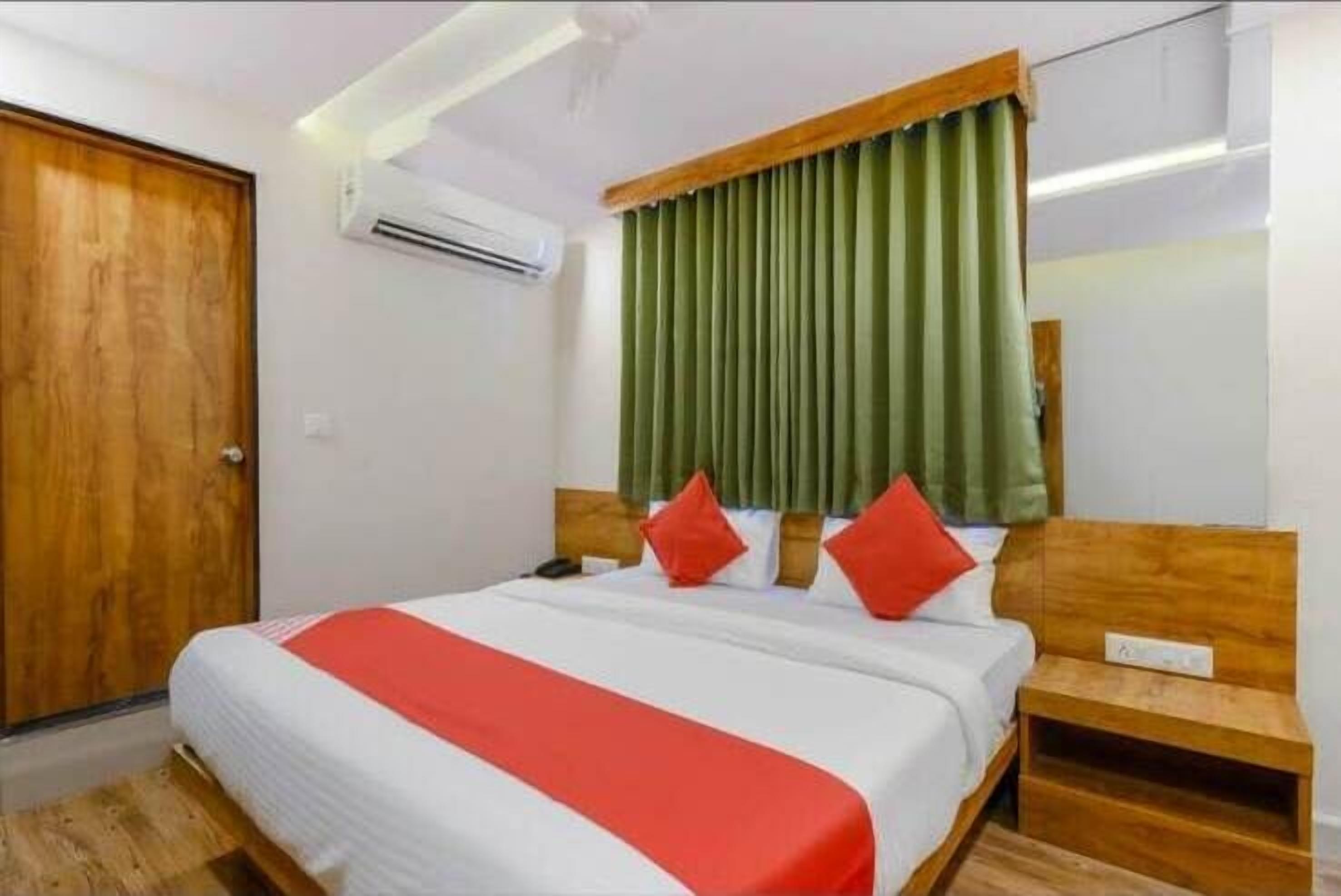Quarto de hotel com porta de madeira, cama com roupa de cama branca e detalhe vermelho, cortinas verdes e cabeceira de madeira.