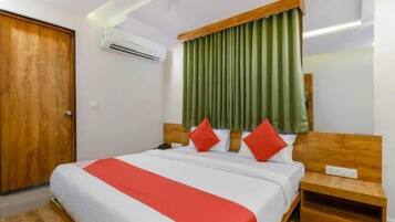 Quarto de hotel com porta de madeira, cama com roupa de cama branca e detalhe vermelho, cortinas verdes e cabeceira de madeira.