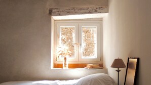 Een slaapkamer met een bed, een raam, een lamp en een vaas met gedroogde planten.