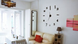 Ein Wohnzimmer mit einem beigefarbenen Sofa, einem Tisch mit einer blumigen Tischdecke, einer großen Wanduhr und einer Lampe.