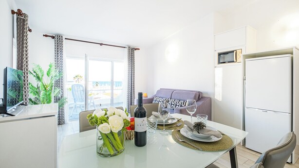 A modern kitchen with a dining table set for two, a refrigerator, and a view of the sea through large windows. - Apartment Atlas (Albufeira)