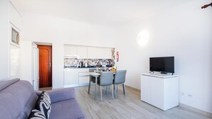 A modern living room with a grey sofa, a dining area with a table and chairs, a kitchen with white cabinets, and a TV on a white stand. - Apartment Atlas (Albufeira)