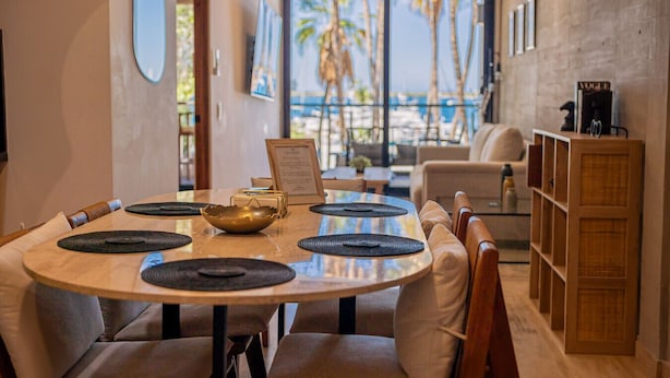 A dining area with a round table, chairs, and a view of palm trees and the ocean through large windows. - 5-Minute Walk to the Malecón — Your Baja Oasis with Sun, Pool & Space to Unwind (La Paz)