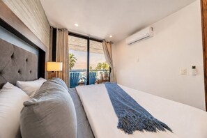 A hotel room with a bed, a nightstand, a lamp, an air conditioning unit, and a view of the outside.