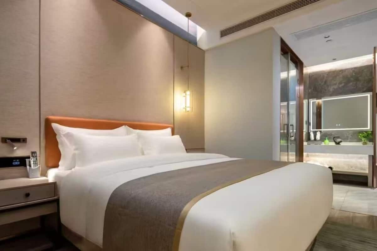 Una habitación de hotel moderna con una cama grande, una mesita de noche con un control remoto y un baño con un espejo amplio y un lavamanos.
