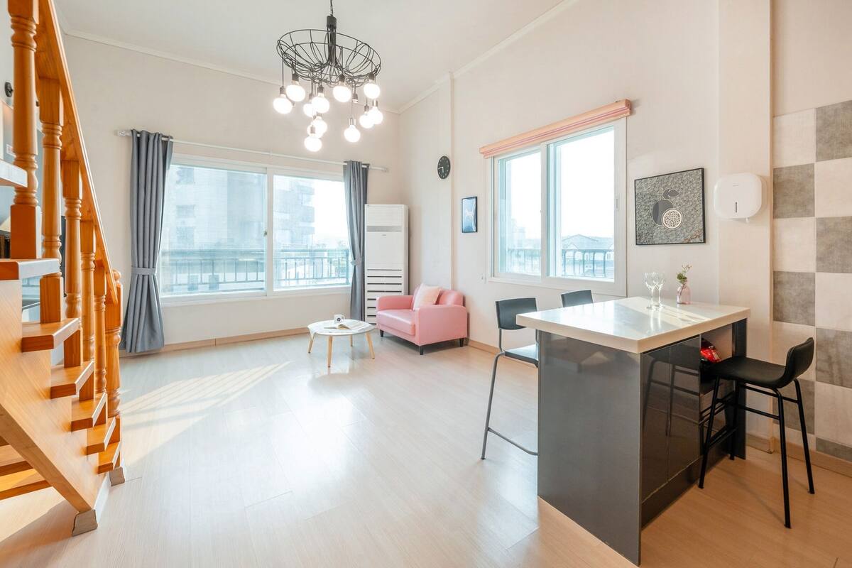 A modern apartment with a kitchen, dining area, and living room. It features large windows, a staircase, and a view of the city.