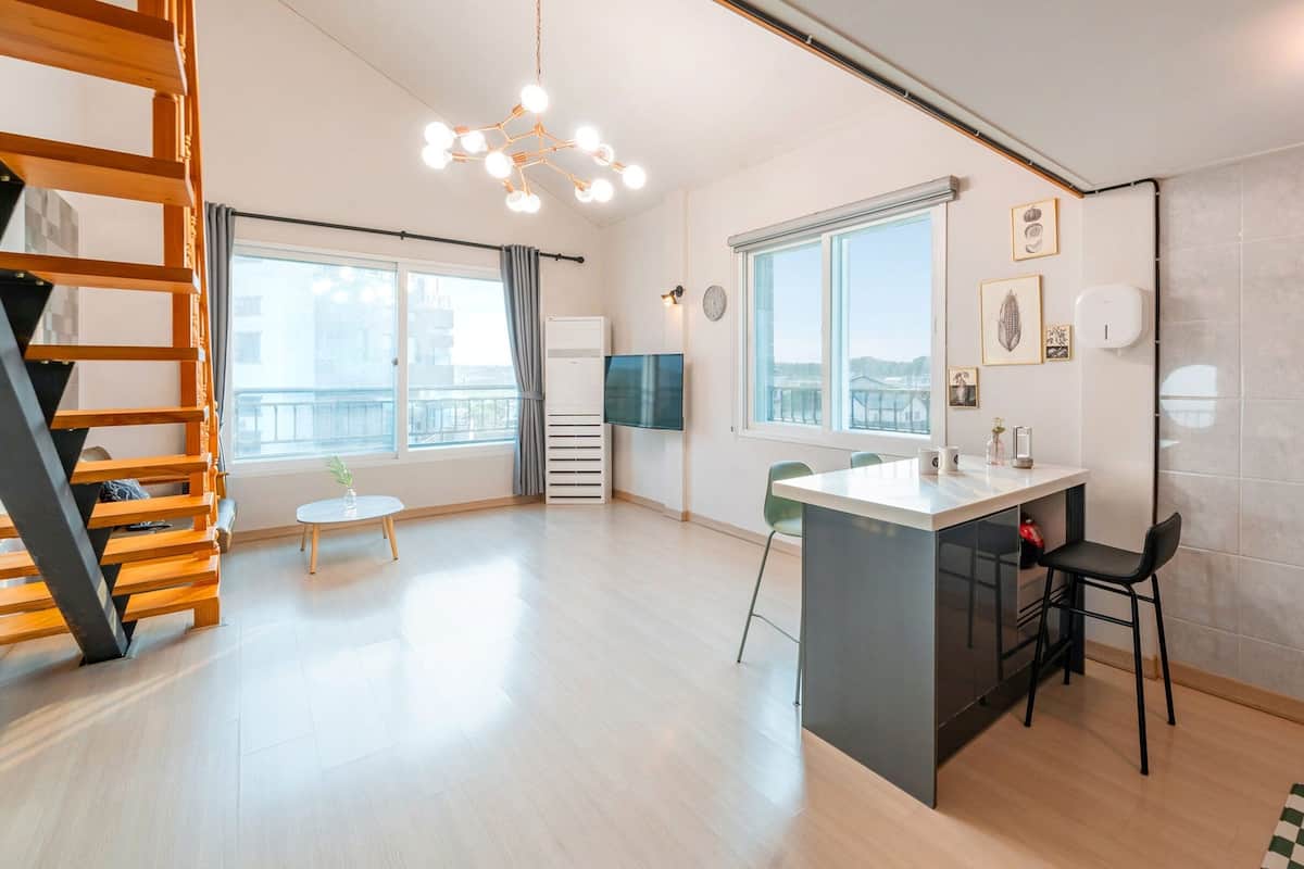 A modern kitchen with a central island, a dining area with chairs, a staircase, and large windows providing natural light.