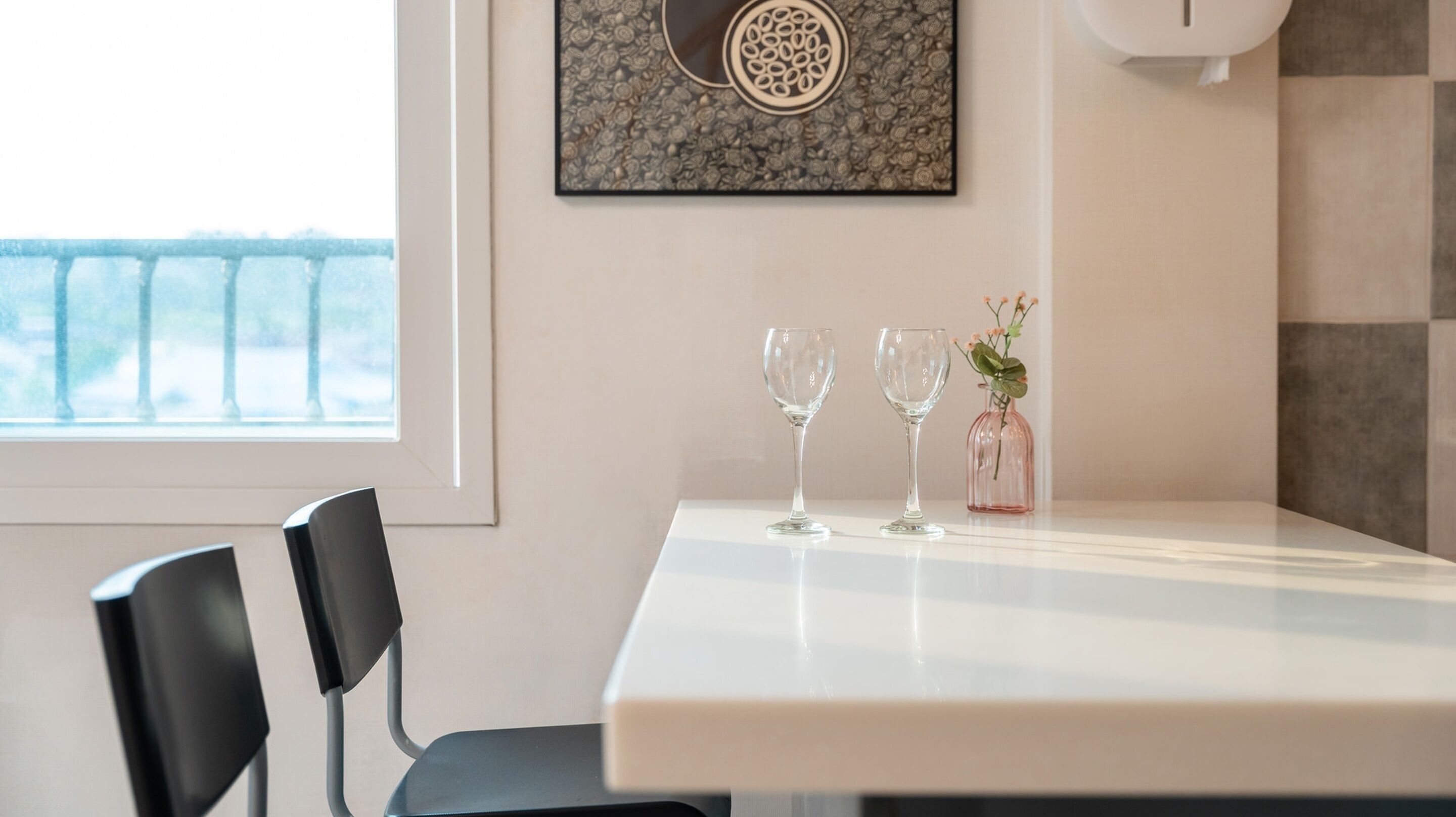 A dining area with a white table, two chairs, two wine glasses, and a small vase with flowers.