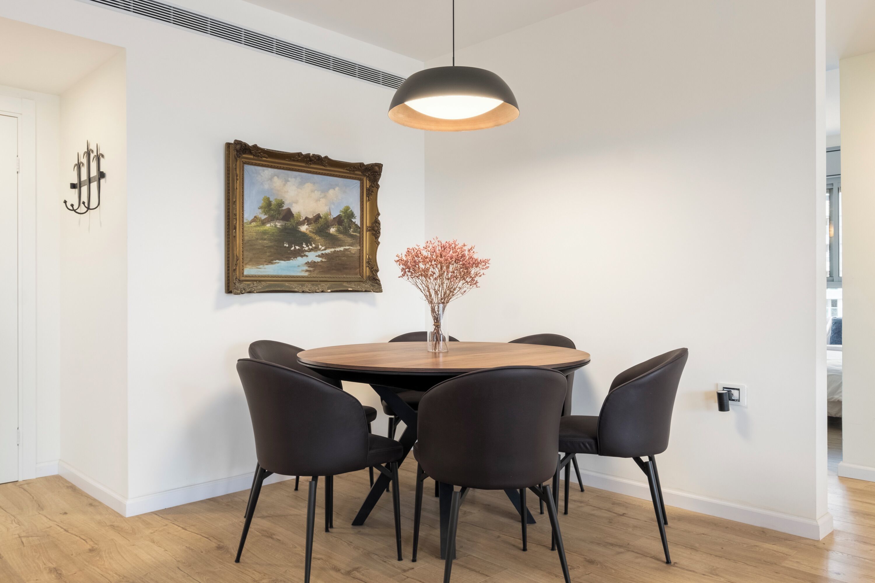 A dining area with a round table, six chairs, a pendant light, a framed painting, and a vase with dried flowers.