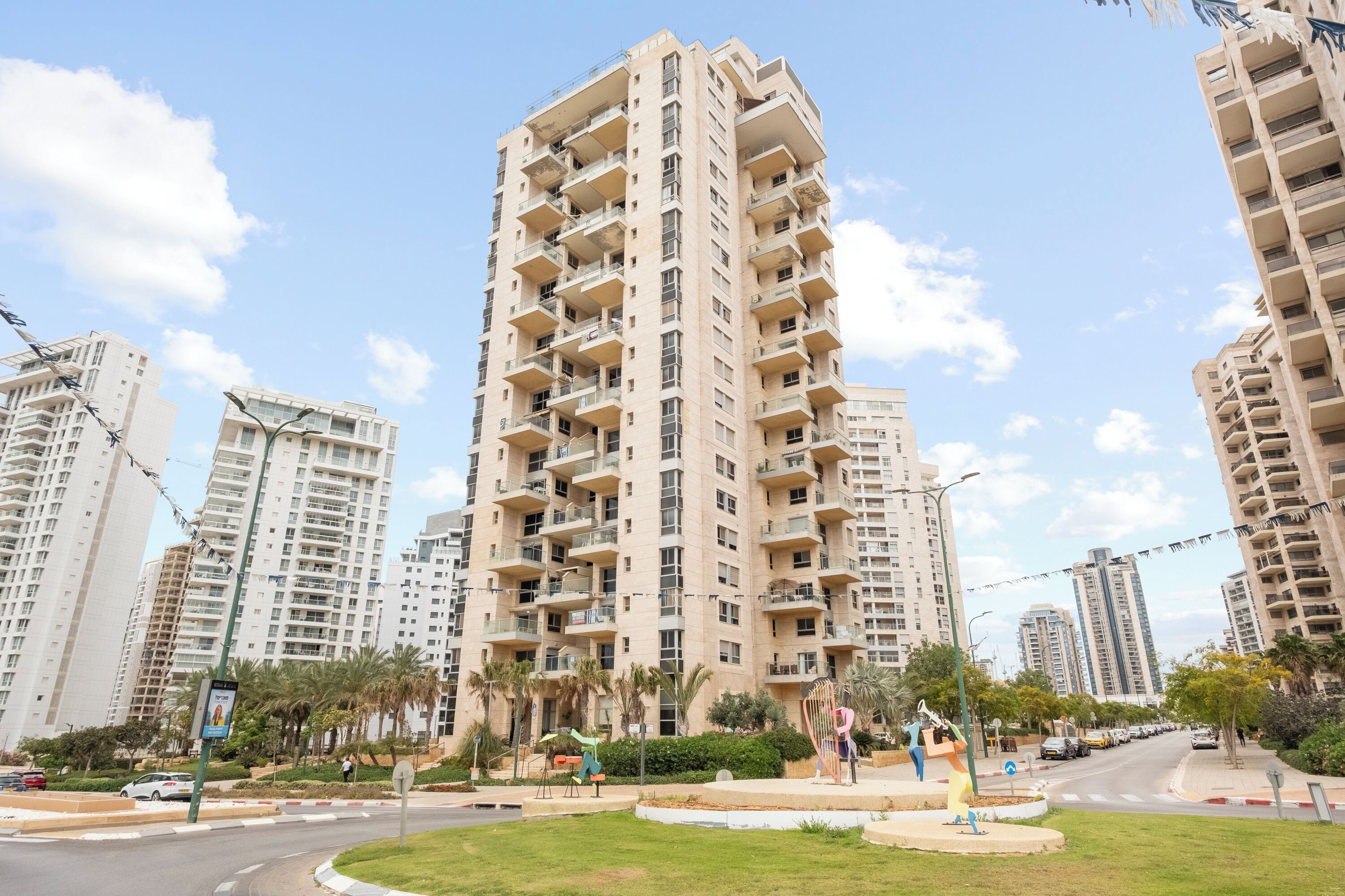 A high-rise residential building with multiple balconies, surrounded by palm trees and a well-maintained lawn.