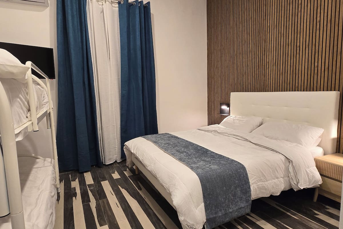 A modern hotel room with a bunk bed, a single bed, a wooden panel wall, a striped floor, and a nightstand with a lamp.