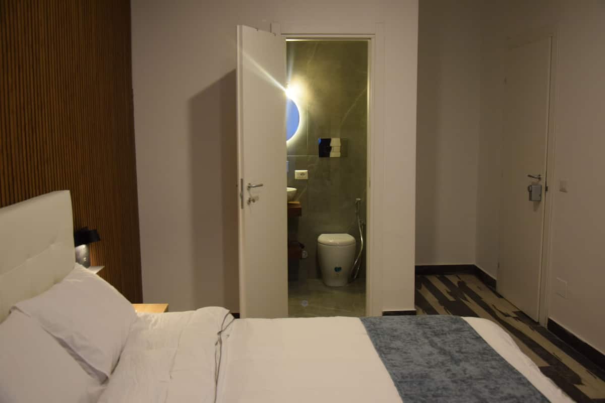 A hotel room with a bed, a nightstand, a wooden panel wall, a bathroom with a toilet and a shower, and a door leading to another room.
