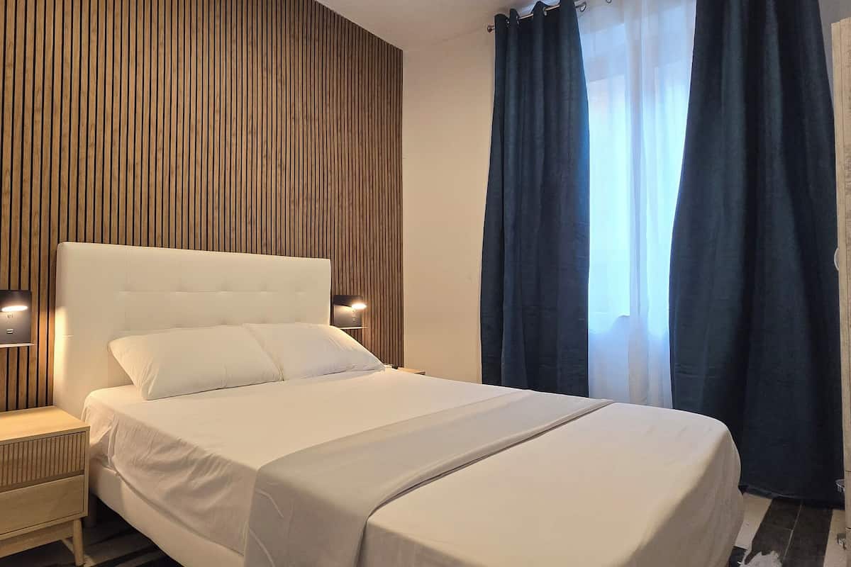 A modern hotel room with a large bed, wooden panel wall, bedside lamps, and a window with curtains.