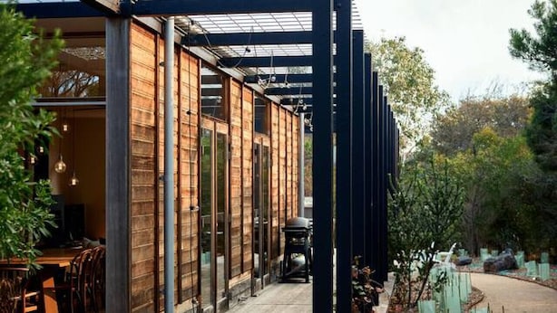 Een modern gebouw met een houten terras en glazen deuren, omgeven door bomen en een grindpad.