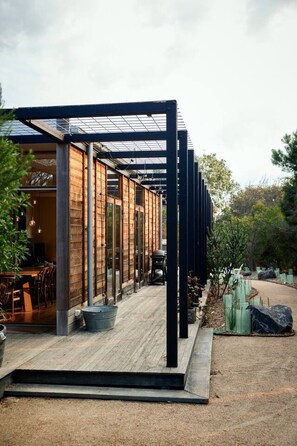 Edifici modern amb terrassa de fusta i portes de vidre, envoltat d'arbres i un camí de grava.