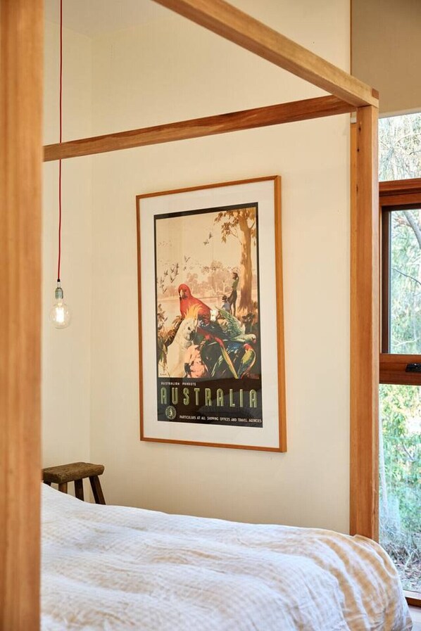 A bedroom with a bed, a framed poster of a parrot, a hanging light, and a window with a view of trees.