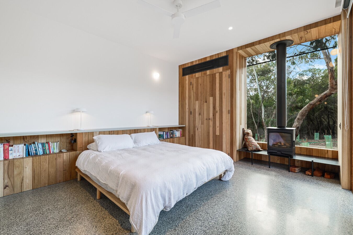 A bedroom with a bed, wooden walls, a built-in bookshelf, a fireplace, and a large window.