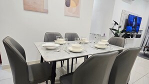 A modern dining area with a rectangular table set for four, surrounded by grey upholstered chairs. There are abstract wall art pieces above the table, and a flat-screen TV mounted on the wall to the right. - Tradres Park 7Pax 3BR2CP (Cheras)