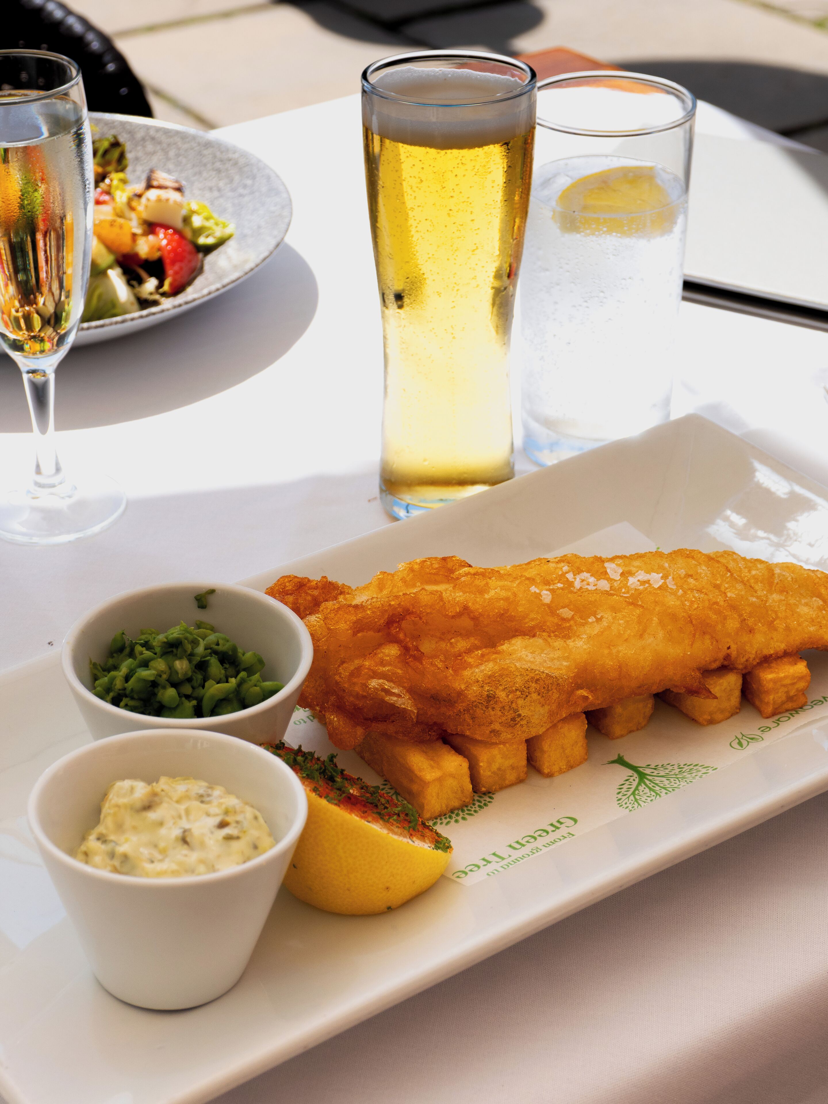 Un plato de pescado y papas fritas, acompañado de guarniciones de guisantes blandos y salsa tártara, junto con una cerveza y una cuña de limón.