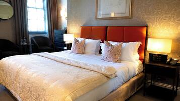 A hotel room with a large bed, two bedside tables, a chair, a desk, a lamp, a framed picture on the wall, and a window with curtains.