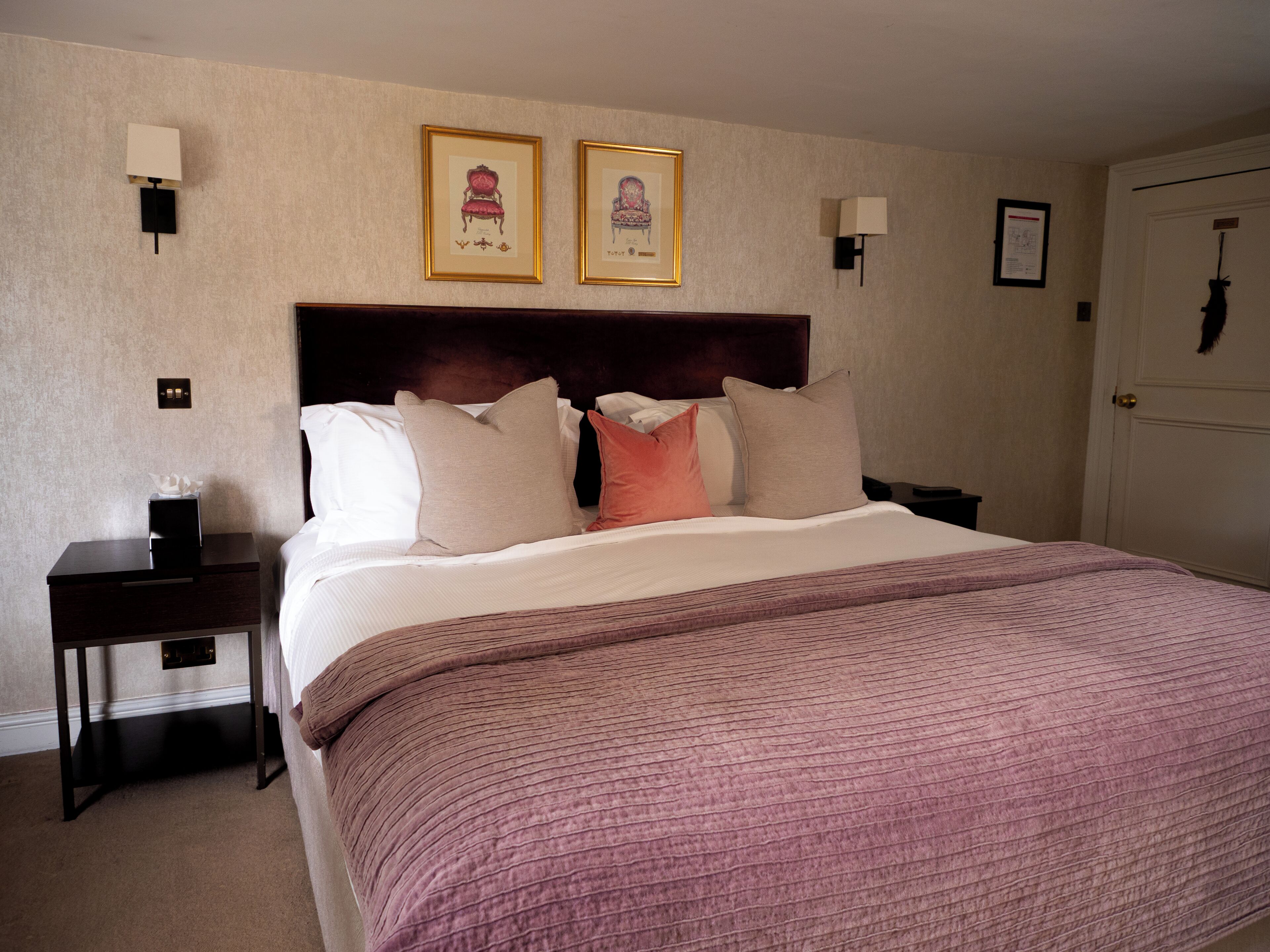 A hotel room with a bed, a nightstand, a headboard, two framed pictures on the wall, and a door.