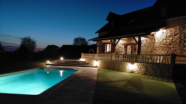 A swimming pool with stone walls and a wooden deck, surrounded by a stone fence and a stone building with a balcony.