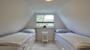 Ett litet, smalt rum med två enkelsängar, ett litet vitt bord och ett fönster med utsikt över träd och hus.