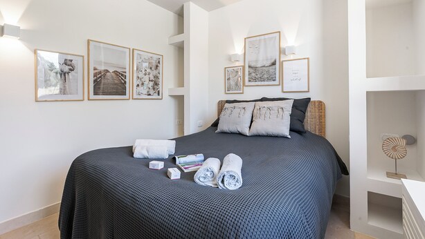Una cama bien hecha con una colcha oscura estampada, dos almohadas y toallas enrolladas. En la pared hay cuadros enmarcados, una mesita de noche con una lámpara y un pequeño estante con un adorno.