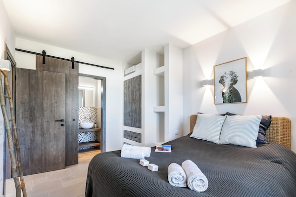 A modern hotel room with a large bed, a wardrobe, a bathroom with a mirror, and a wall-mounted artwork.