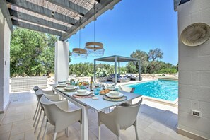 An outdoor dining area with a pool view, white chairs, a table set with plates and glasses, and hanging lights.