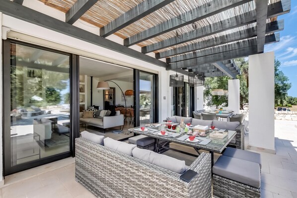 A modern outdoor dining area with a glass enclosure, wicker furniture, and a pergola overhead.