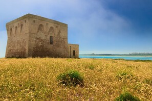 A large, rectangular building with a flat roof and arched windows, situated in a grassy field with a clear blue sky and distant coastline. - Villa Tartaruga Luxury With Pool (Carovigno)