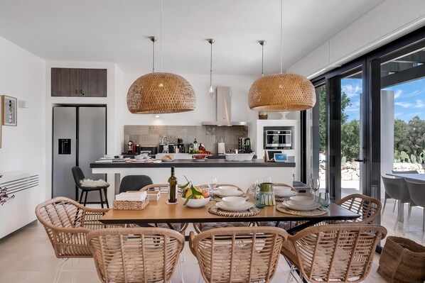 A modern kitchen with a dining area, featuring wicker pendant lights, a large window, and a view of the outdoors. - Villa Tartaruga Luxury With Pool (Carovigno)