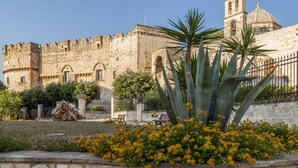 Un edificio histórico de piedra con una torre de campanas y una cúpula, rodeado por un jardín con flores amarillas y una palmera.