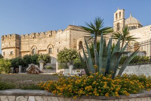 Ein historisches Steinhaus mit Glockenturm und Kuppel, umgeben von einem Garten mit gelben Blumen und einer Palme.