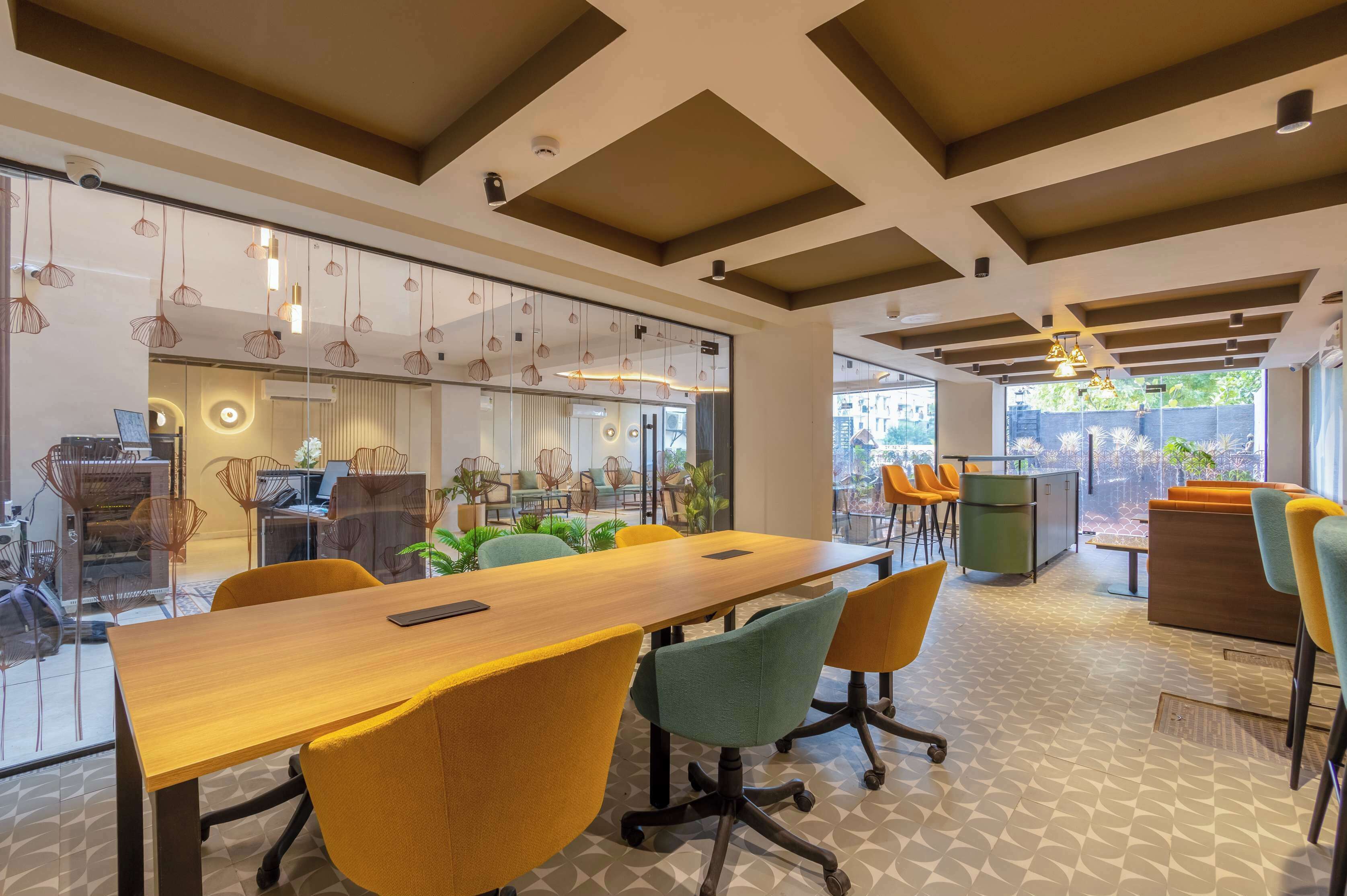 A modern office space with a long wooden table, yellow and green chairs, and a glass partition separating the workspace from a seating area with orange chairs.