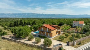 Una casa de dos pisos con techo rojo, piscina y vista a las montañas al fondo.