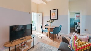 Ein modernes Wohnzimmer mit Flachbildfernseher, einem Essbereich mit rundem Tisch und blauen Stühlen sowie einer gemütlichen Sitzecke mit einem gemusterten Zierkissen.