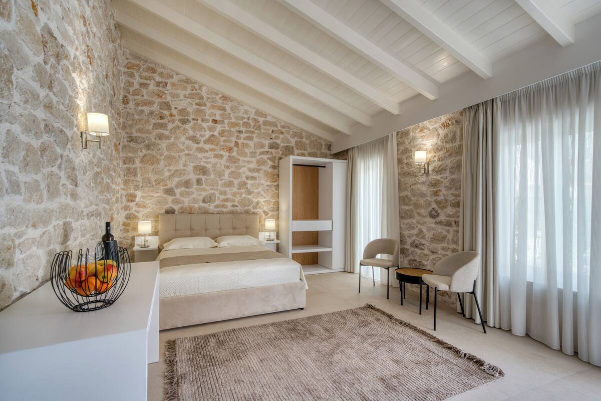 A bedroom with a stone wall, a bed, a nightstand, a chair, and a table.