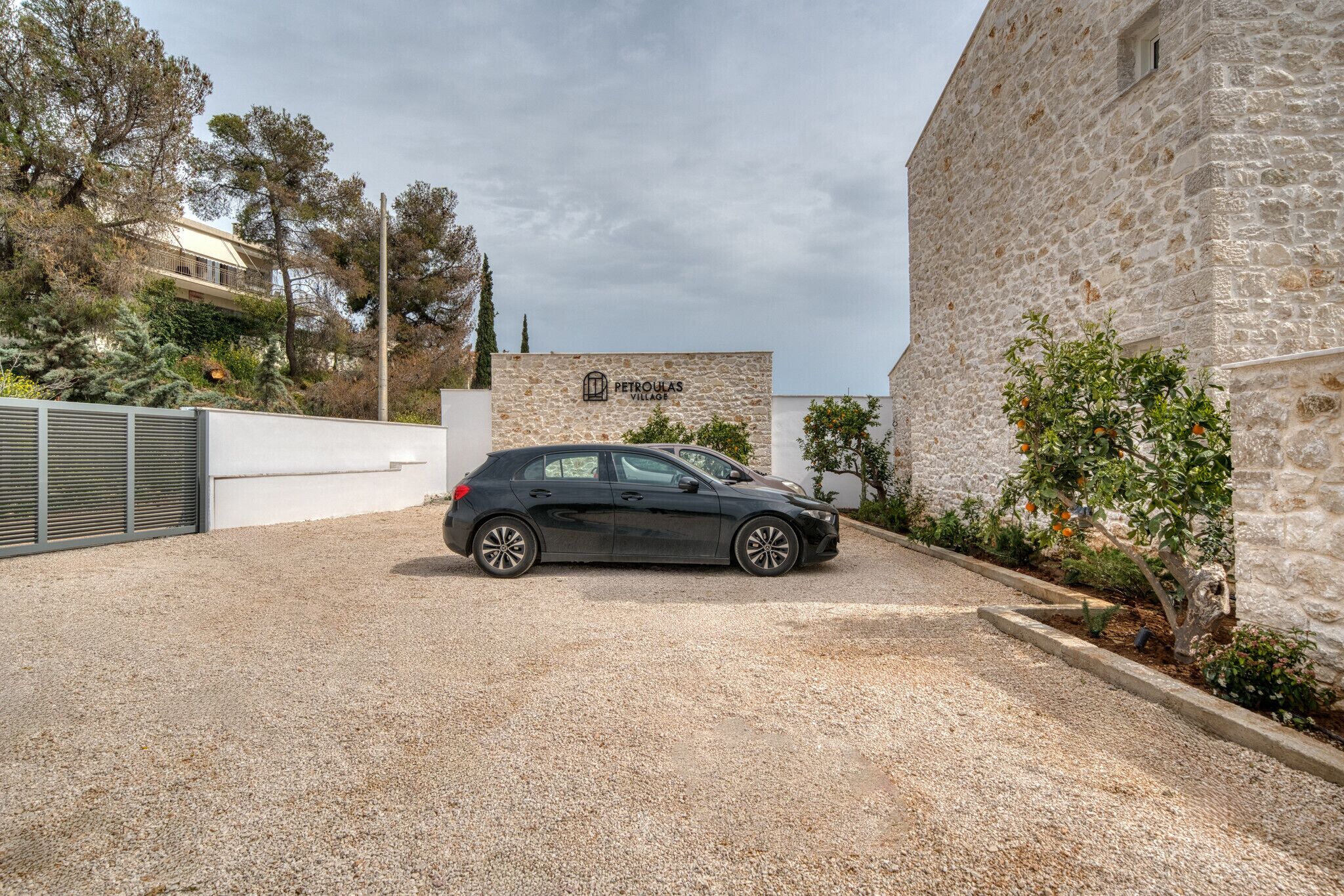 Une voiture noire est garée sur une allée de gravier, et on aperçoit en arrière-plan un bâtiment en pierre sur lequel est inscrit « PETROULAS VILLAGE ».