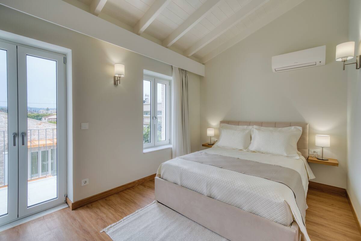 A bedroom with a large bed, two bedside lamps, a window, and an air conditioning unit.