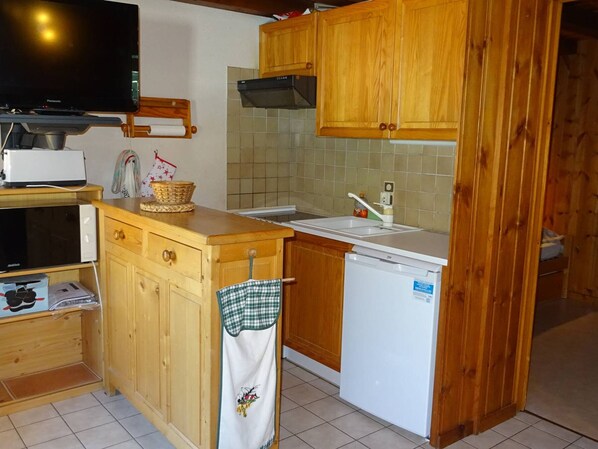 A compact kitchen with wooden cabinets, a built-in microwave, a dishwasher, and a TV mounted on the wall. - Comfy 1BR apt, Wi-Fi, garage, 4 ppl, 150m to shops, 100m to cable cars (Les Gets)