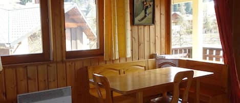 A wooden interior with a dining table and chairs, large windows, and a wall-mounted clock.