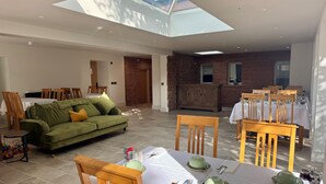 A spacious living area with a high ceiling and skylight, a dining area with wooden chairs and a table set for two, and a cozy green sofa with cushions. - Redstones Hotel (Appleby-in-Westmorland)