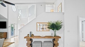 Ein modernes Wohnzimmer mit einem Holzkonsolentisch, zwei Poufs, einer großen Topfpflanze und einem gerahmten Schild an der Wand.