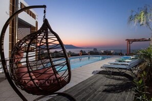 A poolside area with a hanging chair, lounge chairs, and a view of the ocean at sunset.
