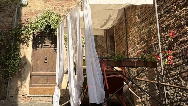 A narrow street with a building featuring a wooden door and a window with shutters. Clothes are hanging on a line outside the building. - Pendulum - 9 (Siena)