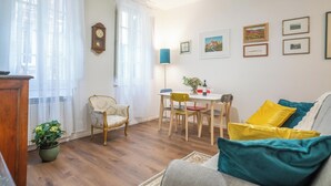 A living room with a wooden floor, a dining table with chairs, a sofa with cushions, a potted plant, and a wall clock. - Pendulum - 9 (Siena)